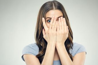 Eine Person guckt in die Kamera, hat beide Hände vor dem Gesicht und guckt mit einem Auge durch die Hand durch